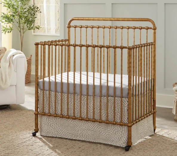 Golden metal crib with mattress and patterned skirt. Set on a carpeted floor, near a white chair.