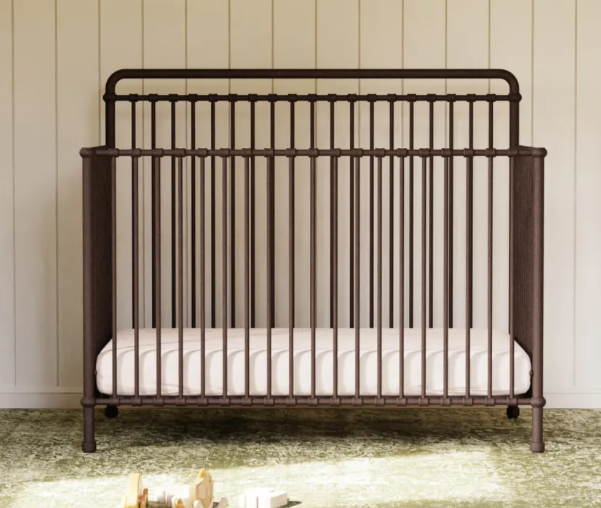 Brown metal crib with vertical bars, white mattress, against a light wall, on a green rug.