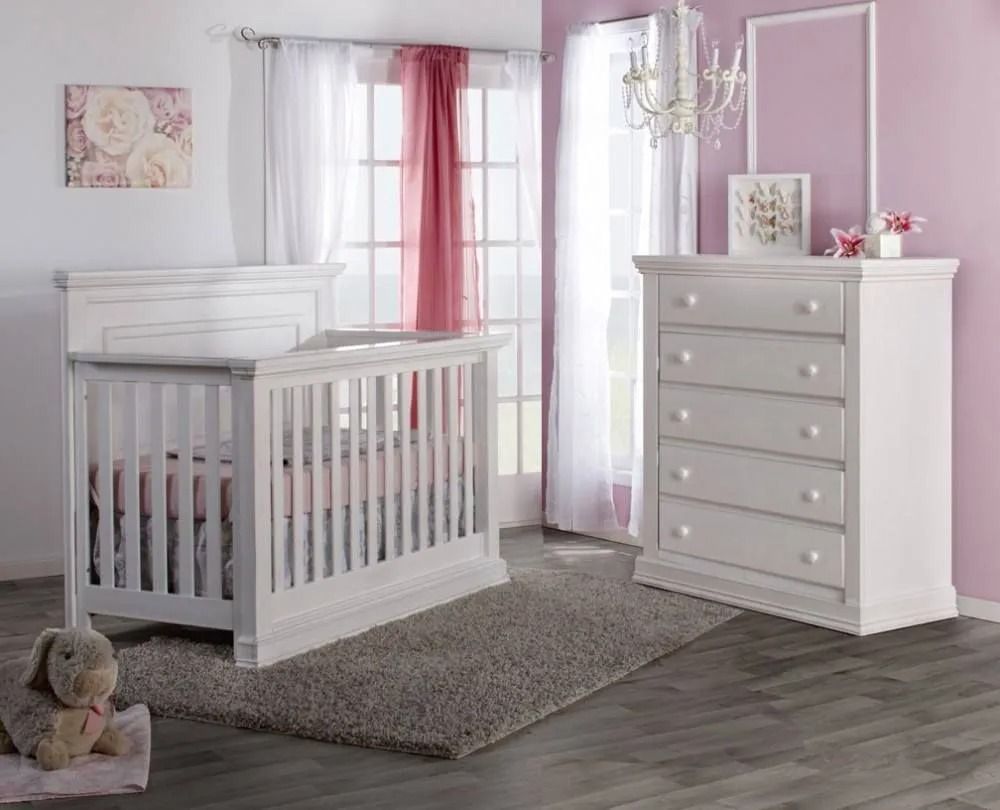 White nursery furniture set: crib, dresser, rug, window, pink curtains, artwork.