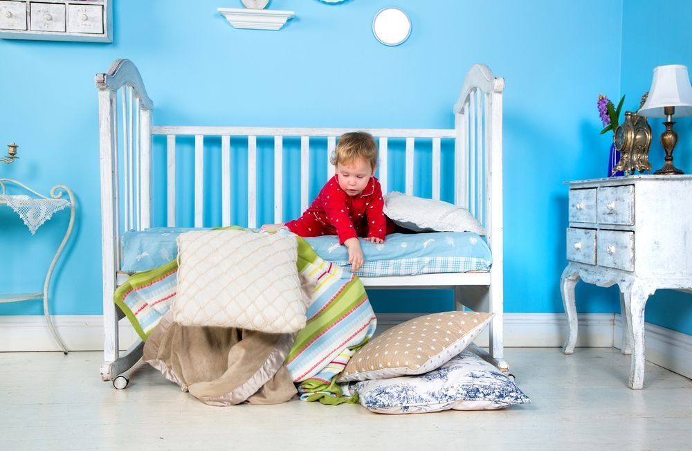 Child in red pajamas playing with pillows and blankets in a white crib, blue room.