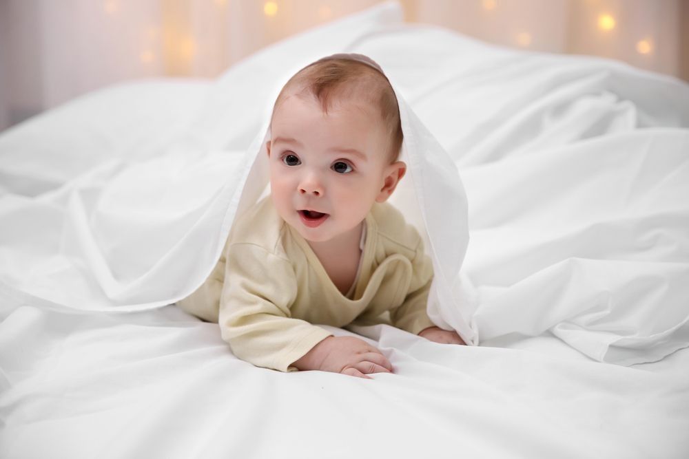 Baby peeking from under white blanket on a bed, light-colored clothing, joyful expression.