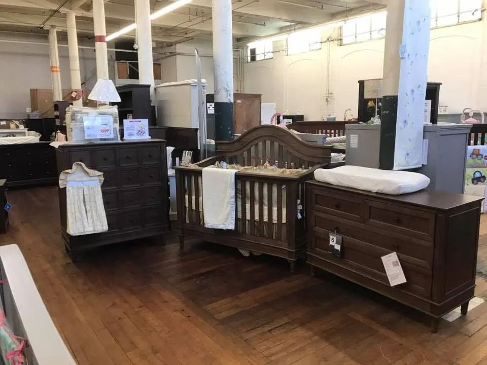 Baby furniture set in a showroom: crib, dresser with changing pad, and chest of drawers; wood floors and white columns.