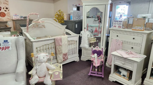 Baby furniture display in a store, featuring a crib, dresser, and armchair. Light colors, soft toys, and decor.