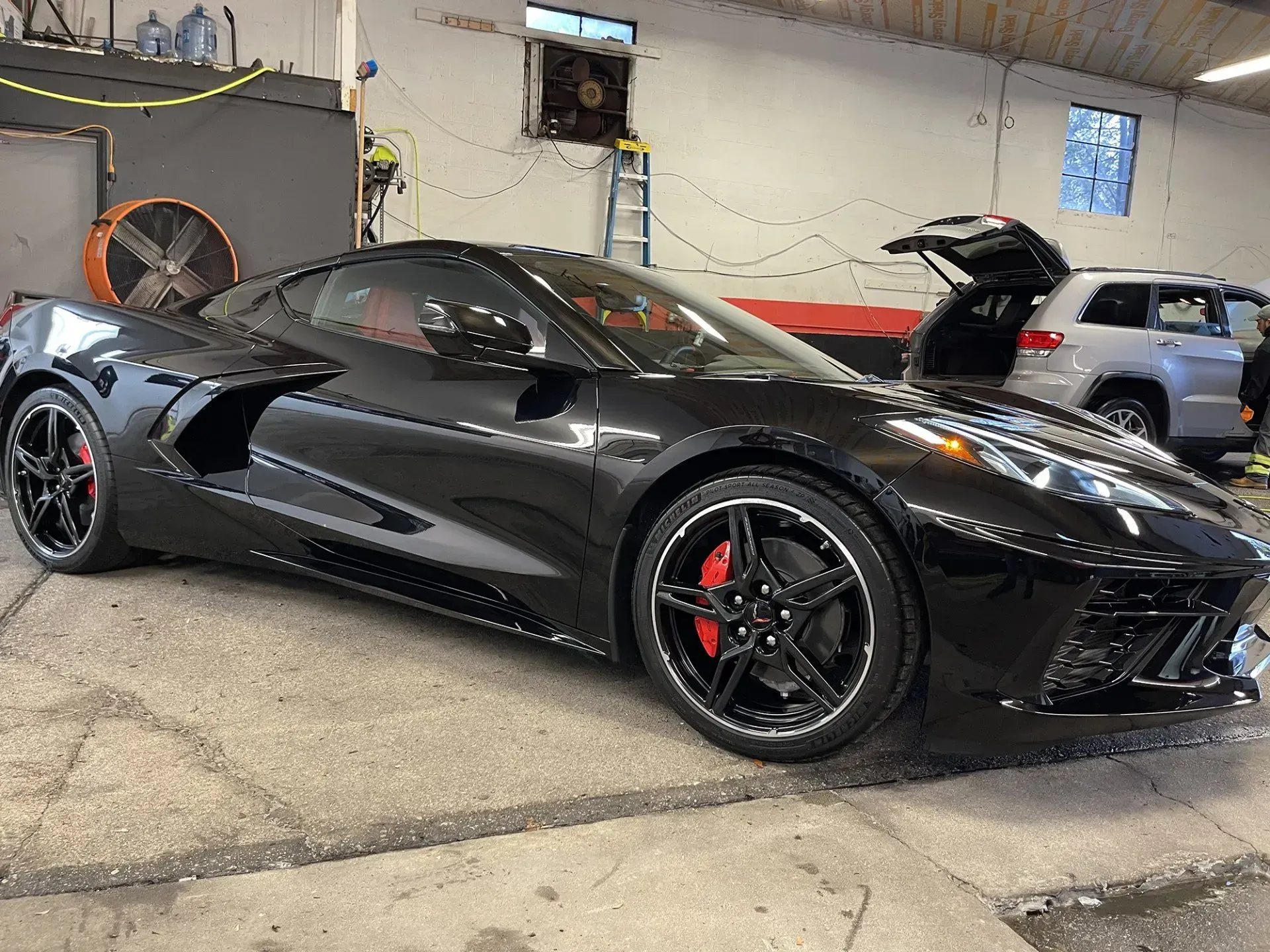 Black Corvette sports car with red brake calipers, parked inside a garage, next to a gray SUV.