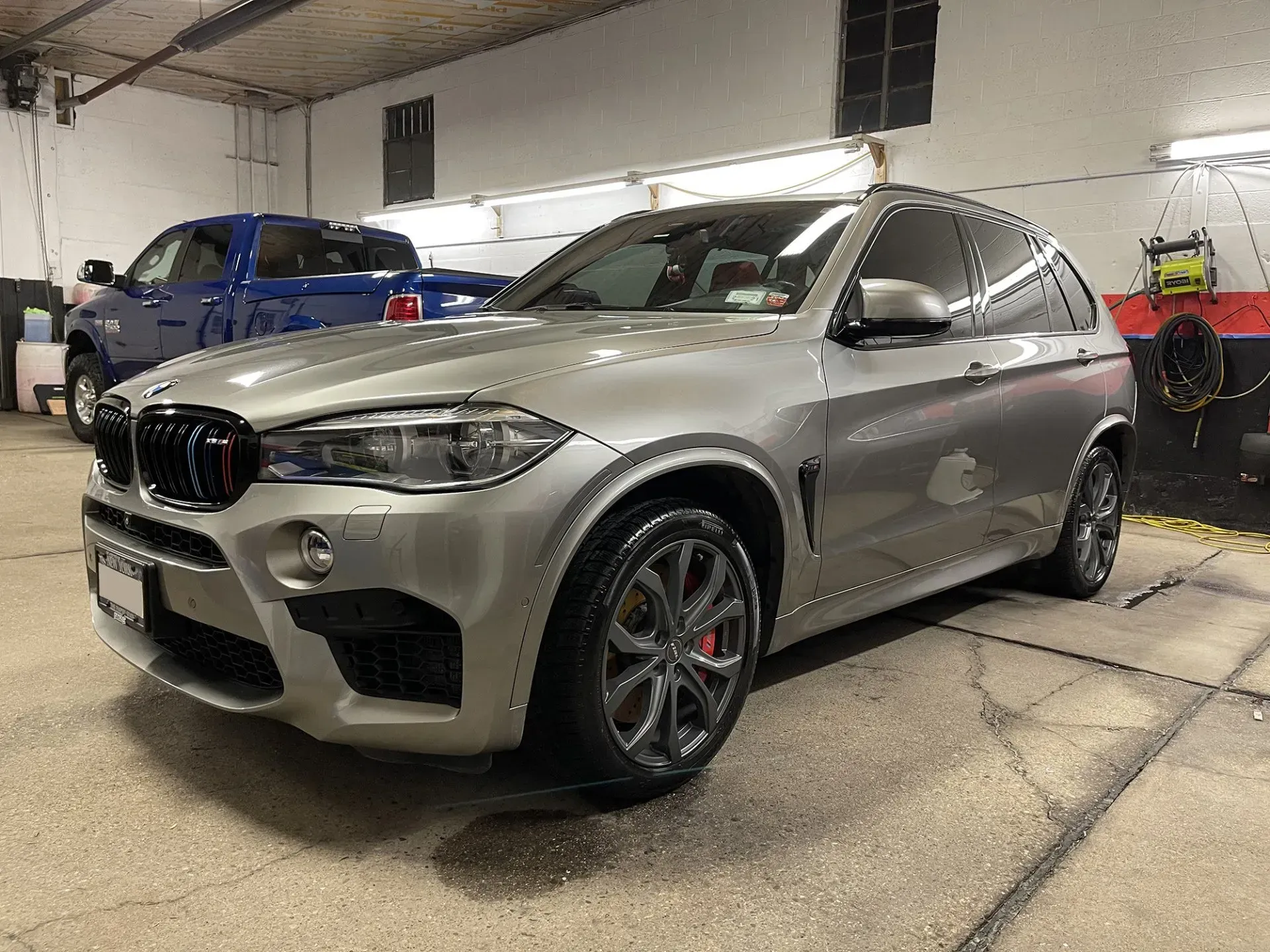 Silver BMW X5 M SUV parked inside a garage, with a blue pickup truck visible in the background.