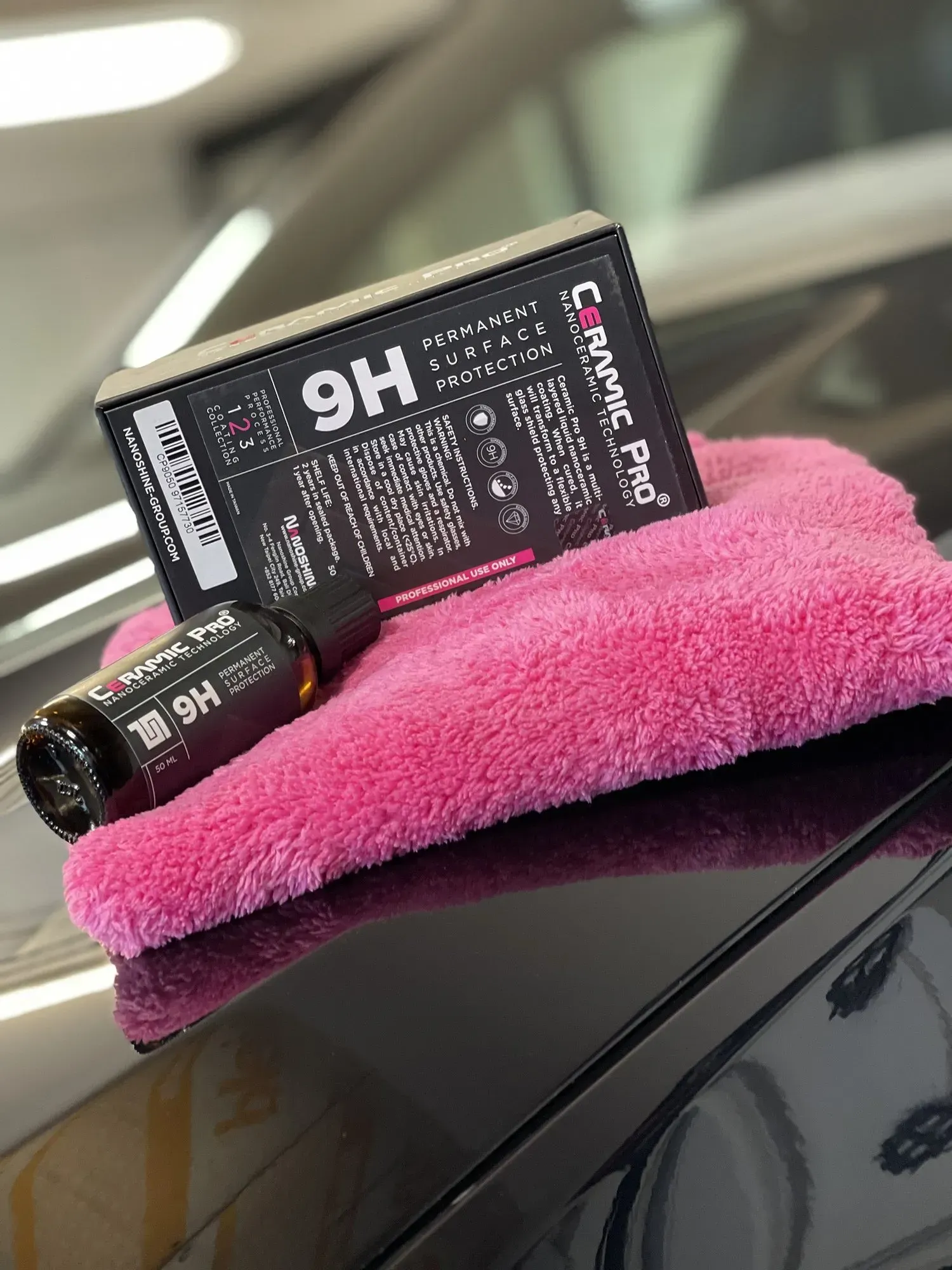 Black bottle and box of Ceramic Pro 9H coating on pink microfiber towel, sitting on a car's black surface.