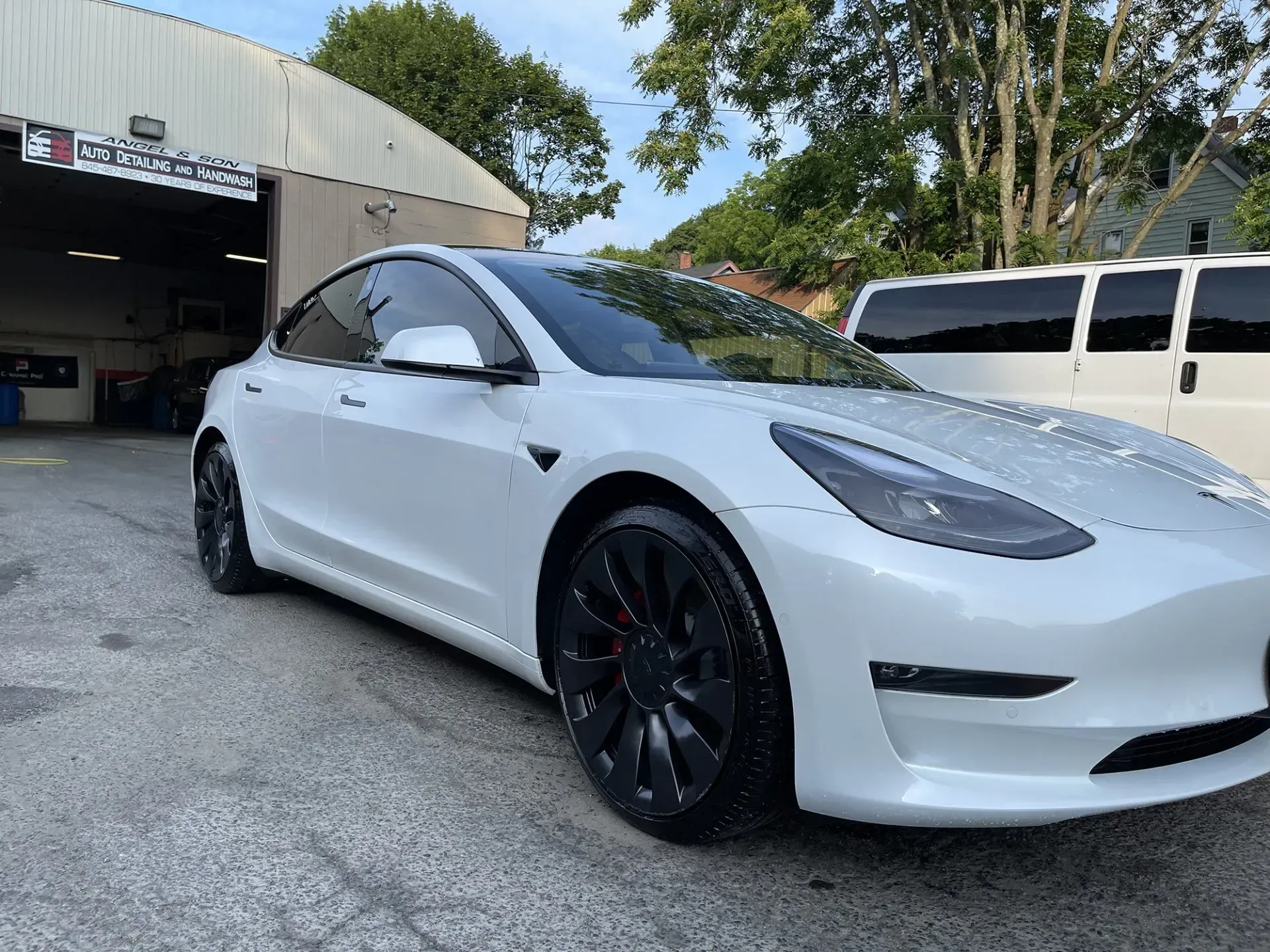 White Tesla parked in front of a shop with black wheels; a van is behind it.