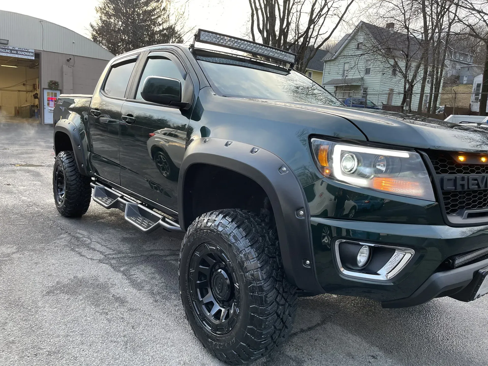Dark green lifted Chevy Colorado pickup truck with black wheels, light bar, and fender flares.
