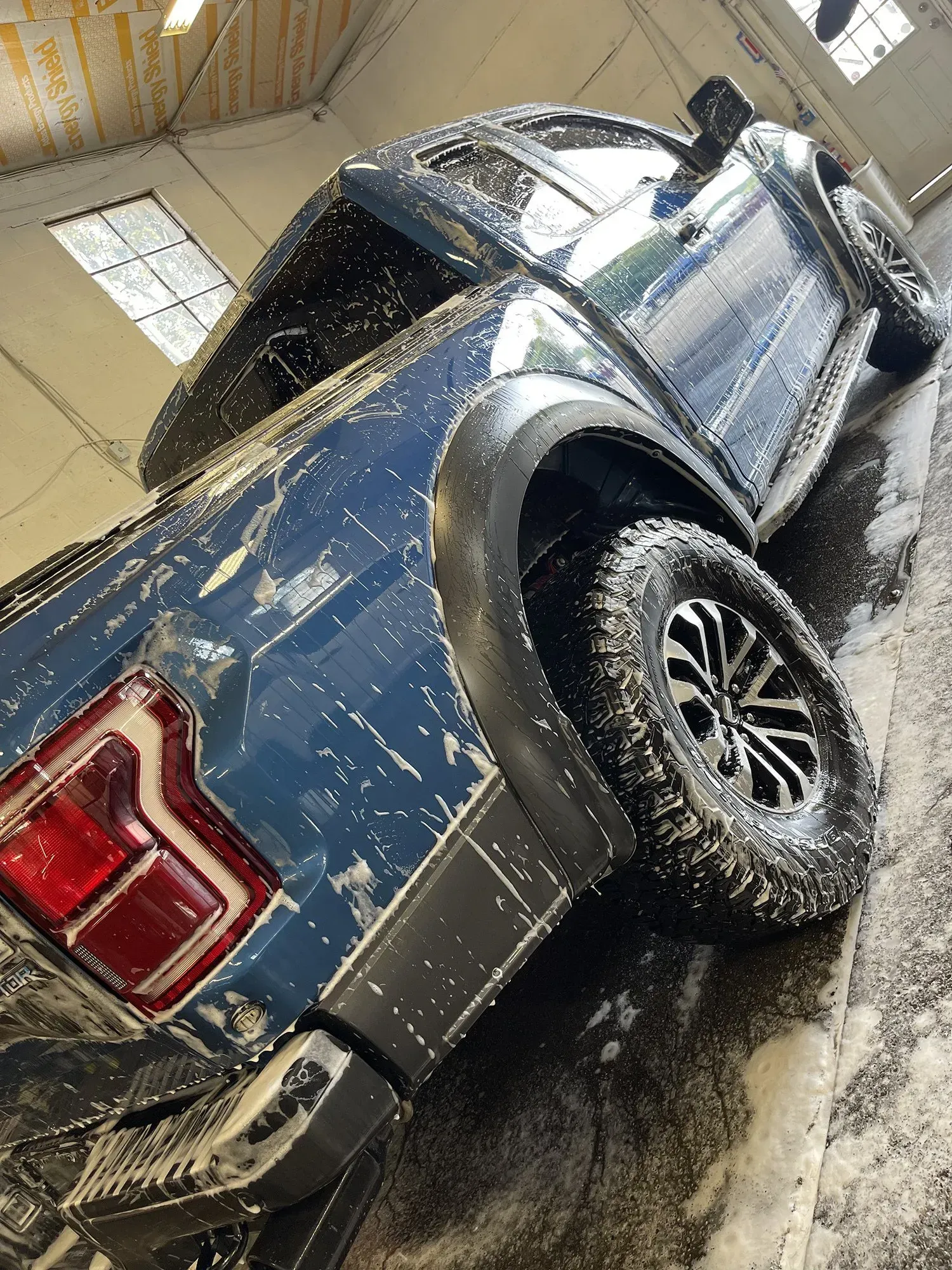 Blue Ford Raptor truck covered in foam, being washed indoors.