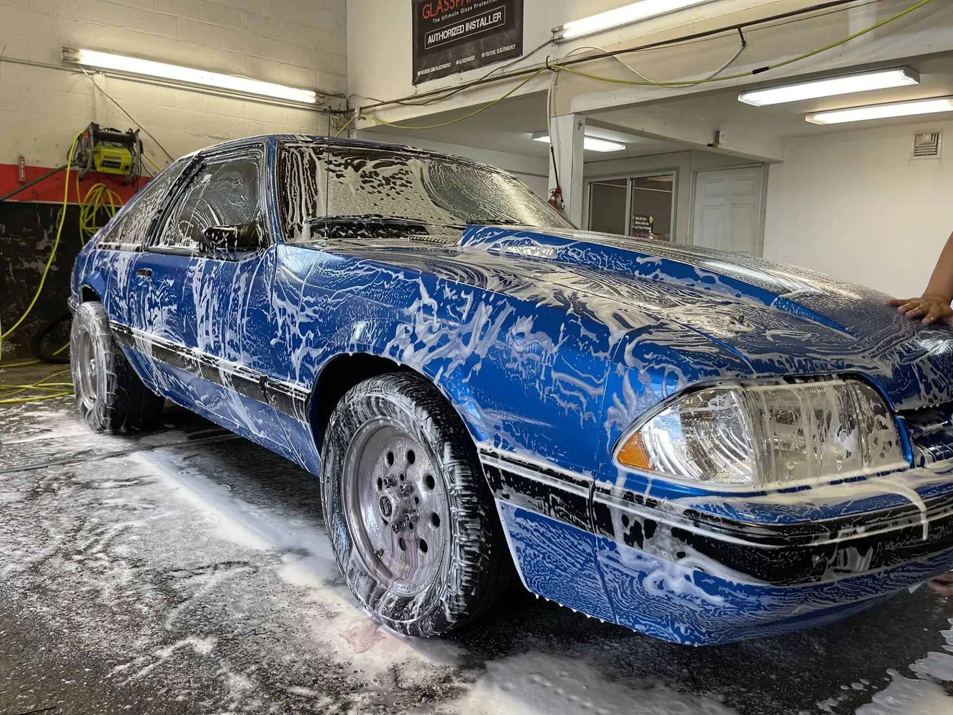 Blue Ford Mustang covered in soap suds being washed in a shop.
