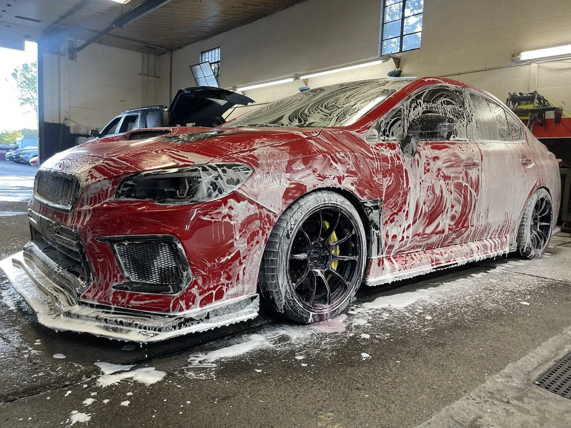Red Subaru being washed with soapy foam in a carwash setting.