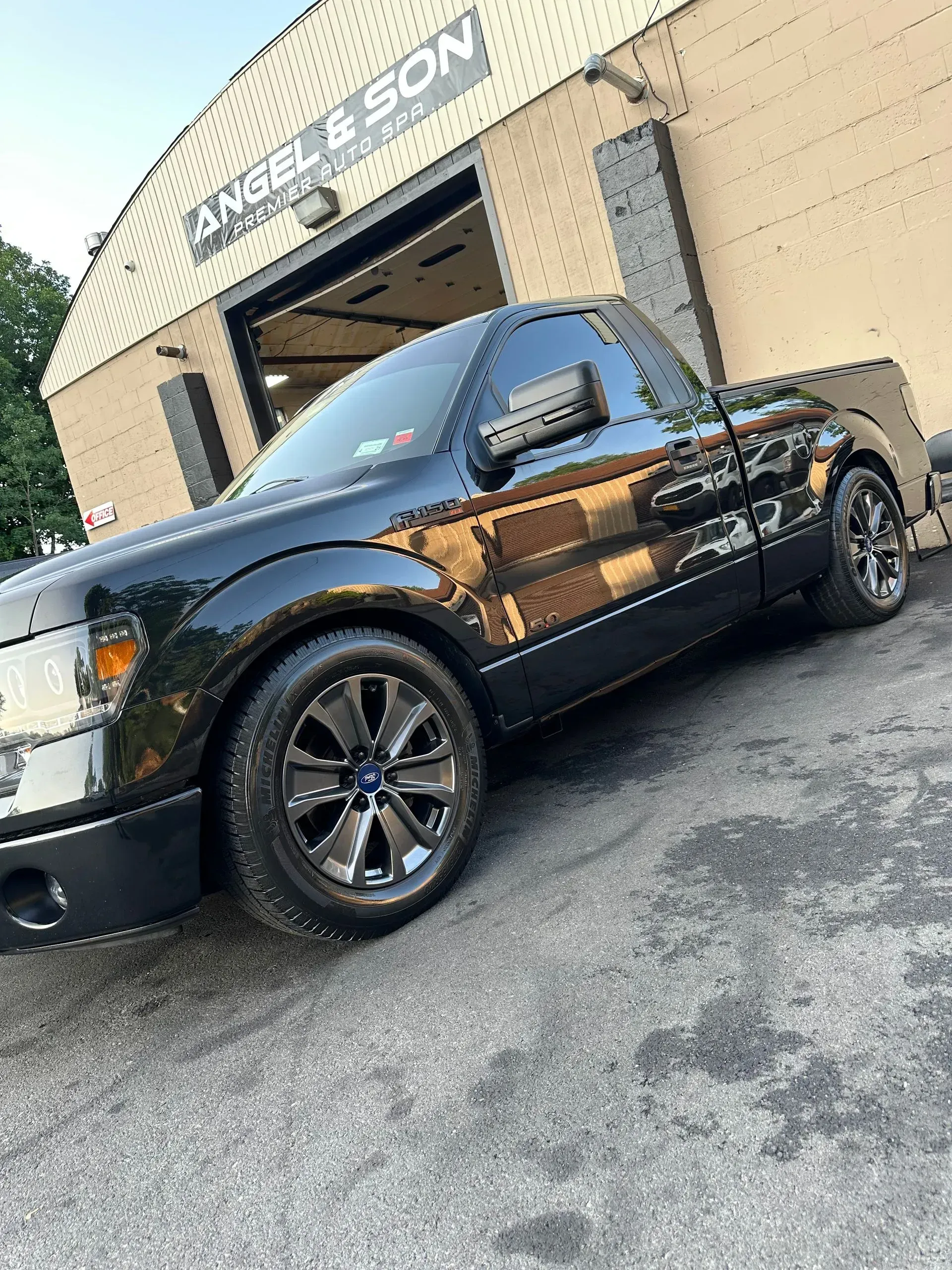 Black Ford F-150 pickup truck with tinted windows and dark rims parked in front of a repair shop.