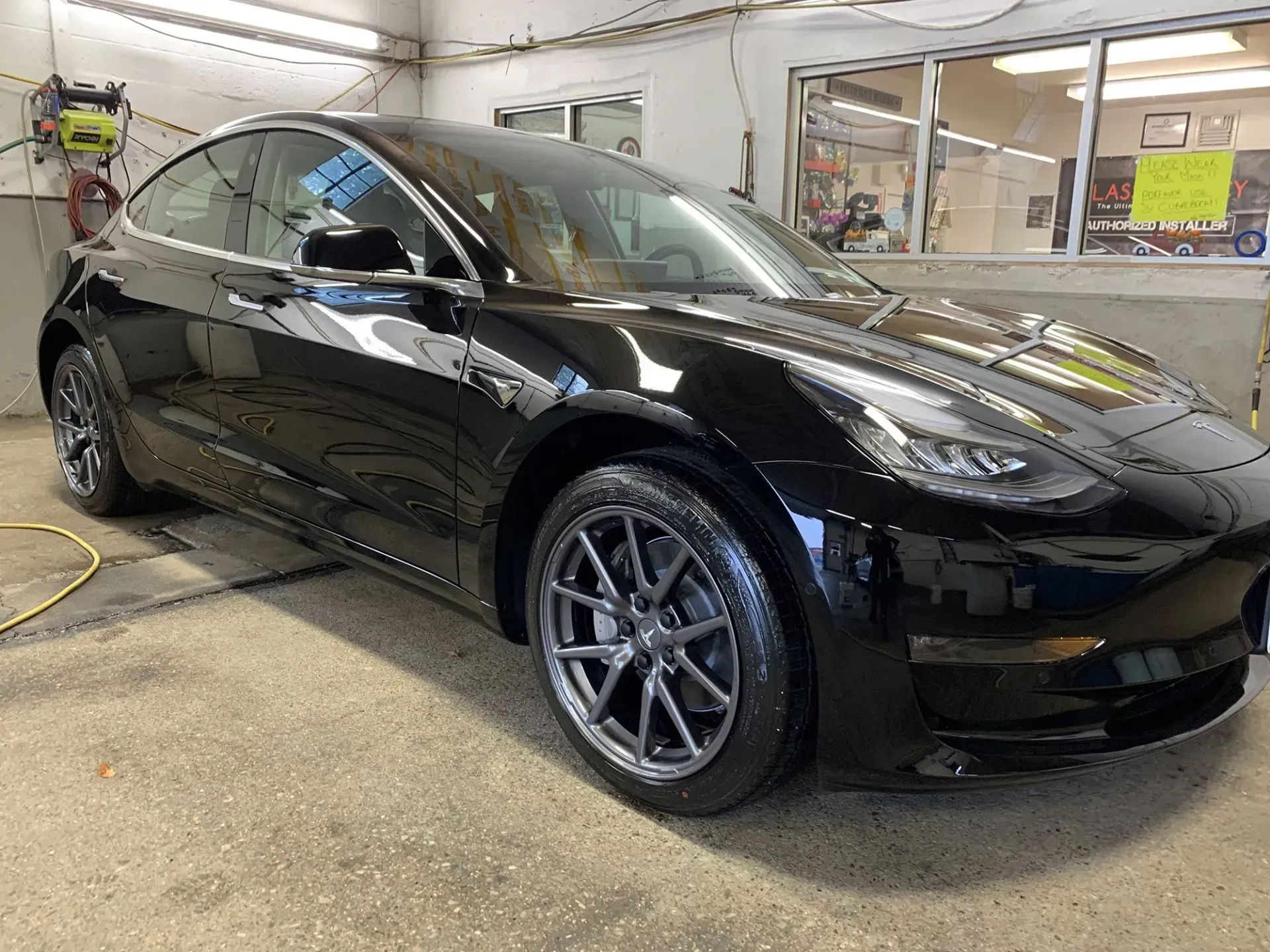 Black Tesla Model 3 car at a car wash, glossy paint, grey wheels, bright indoor lighting.