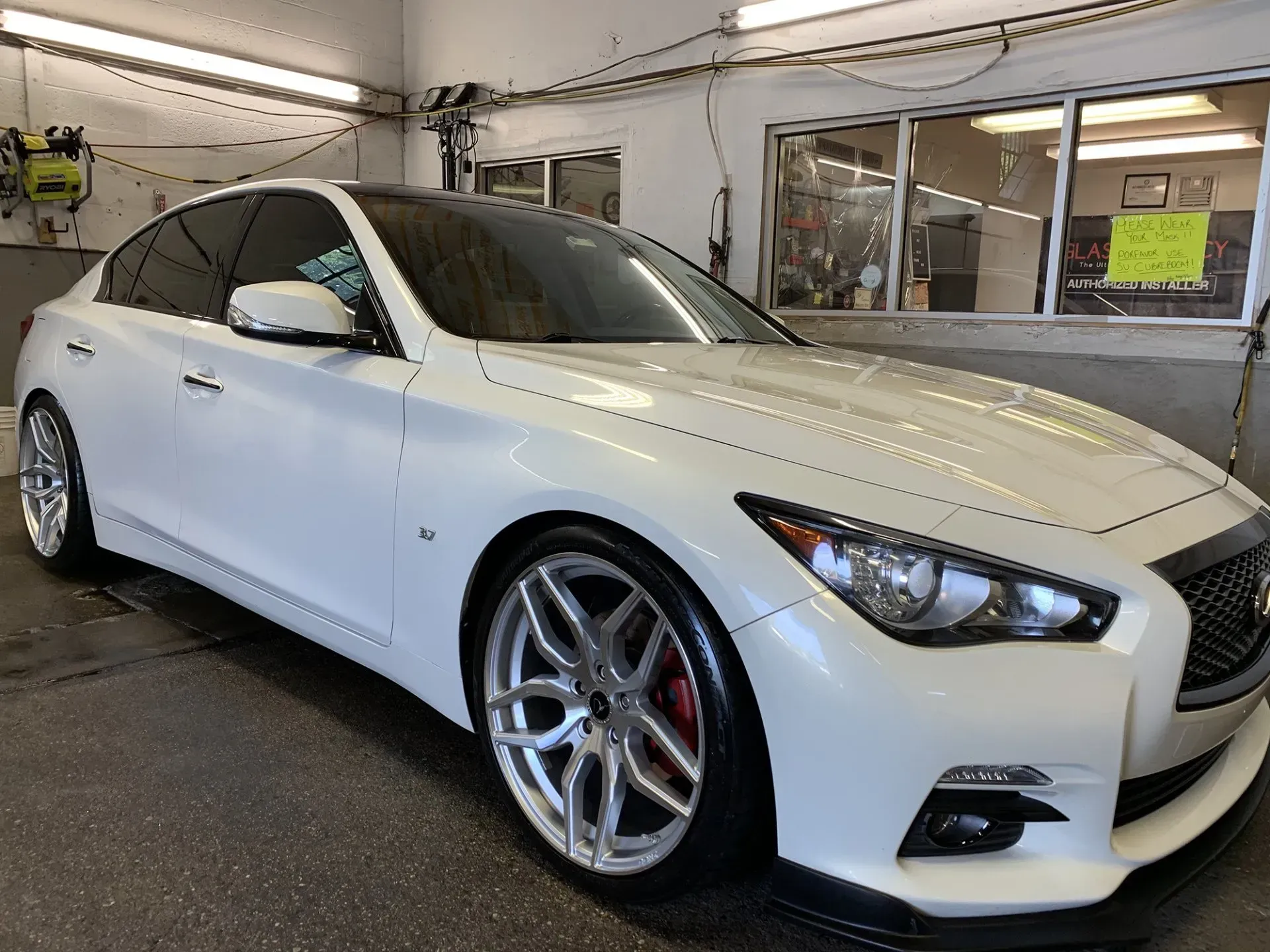 White Infiniti Q50 with tinted windows, custom rims, and red brake calipers, parked inside a car wash bay.