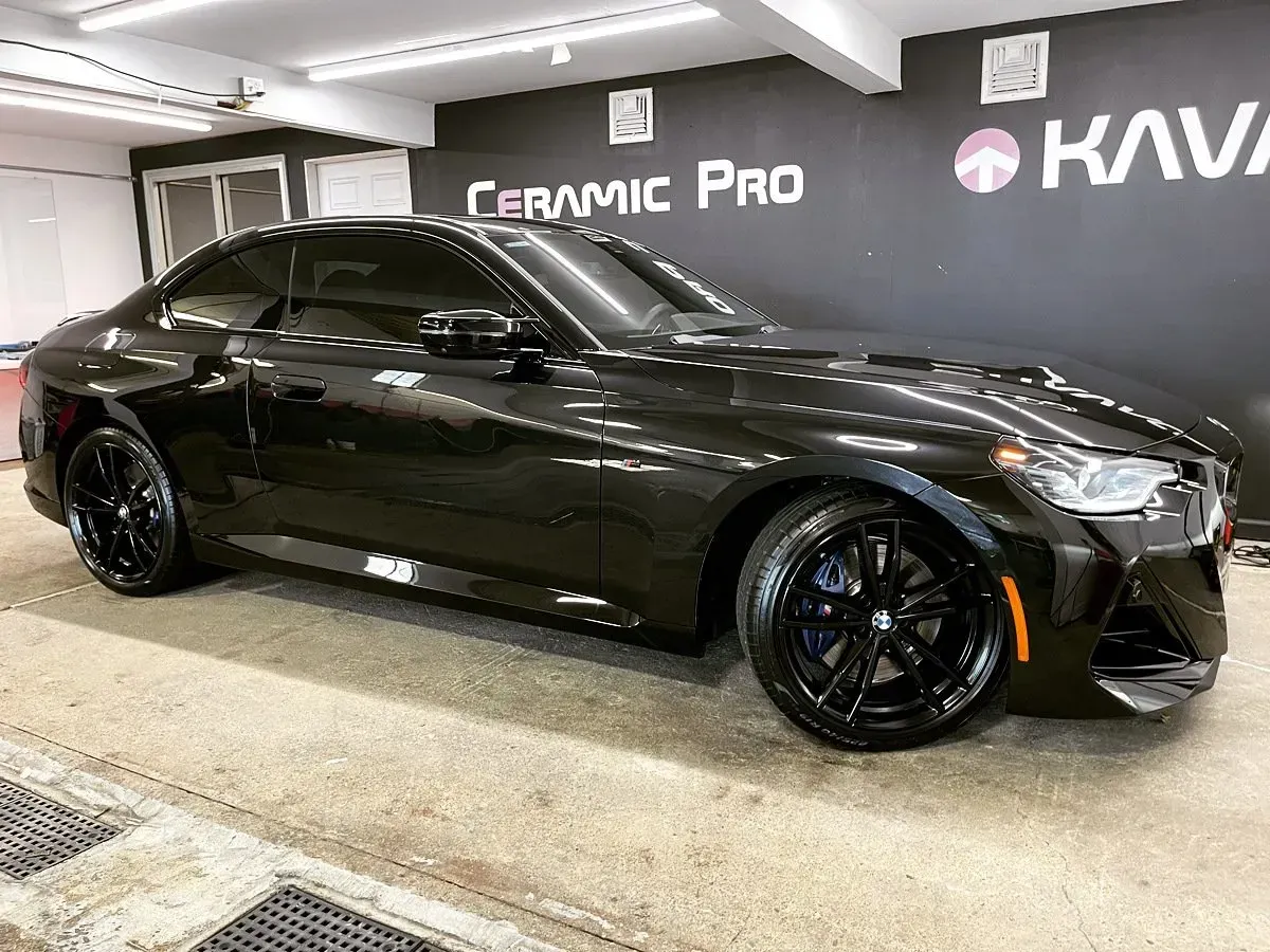 Black BMW coupe, freshly detailed, parked inside a garage. Glossy paint, black wheels, Ceramic Pro sign in background.