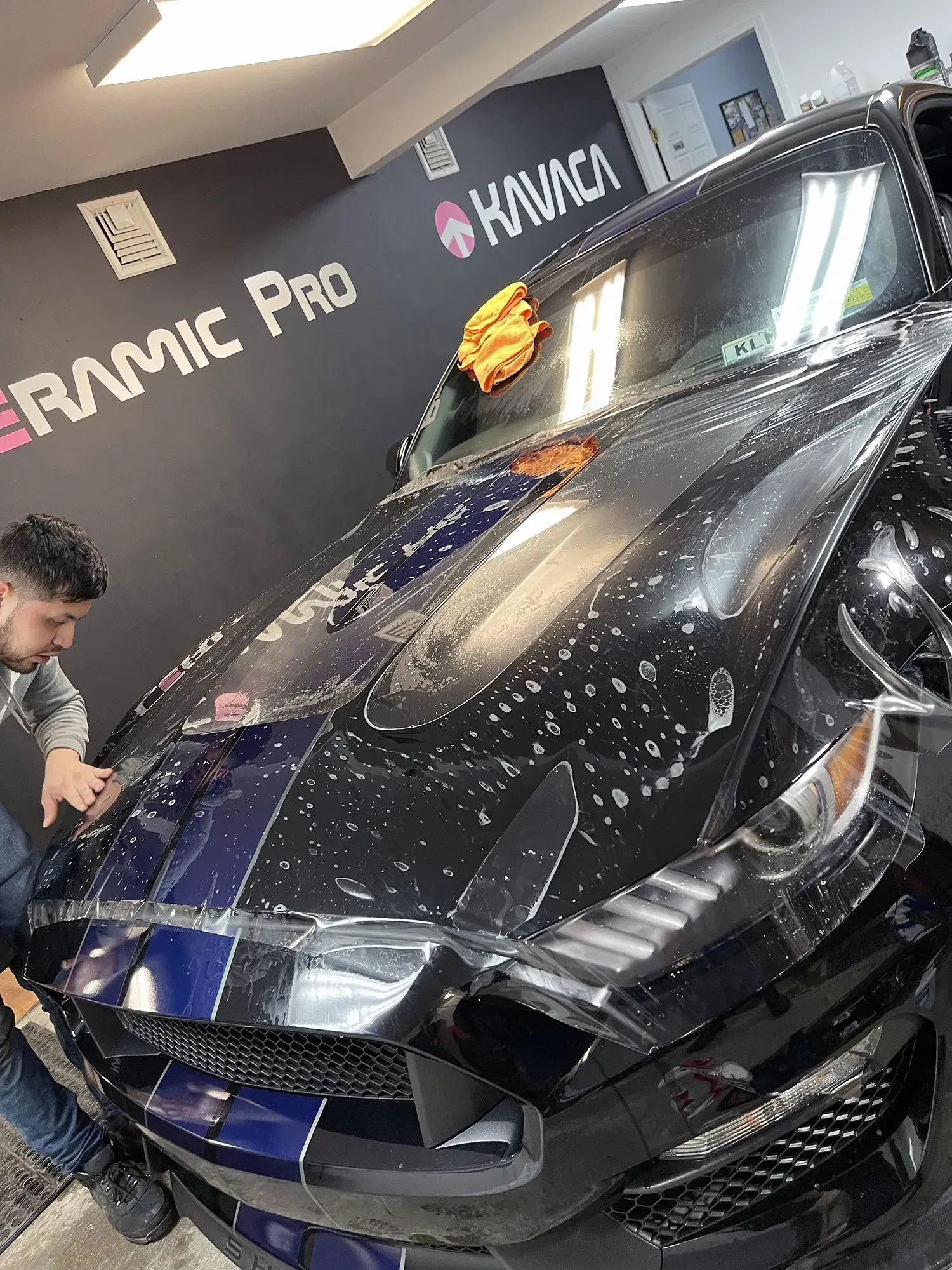 Man applying film to black Mustang hood in a shop; blue stripes, bubbles, orange cloth.