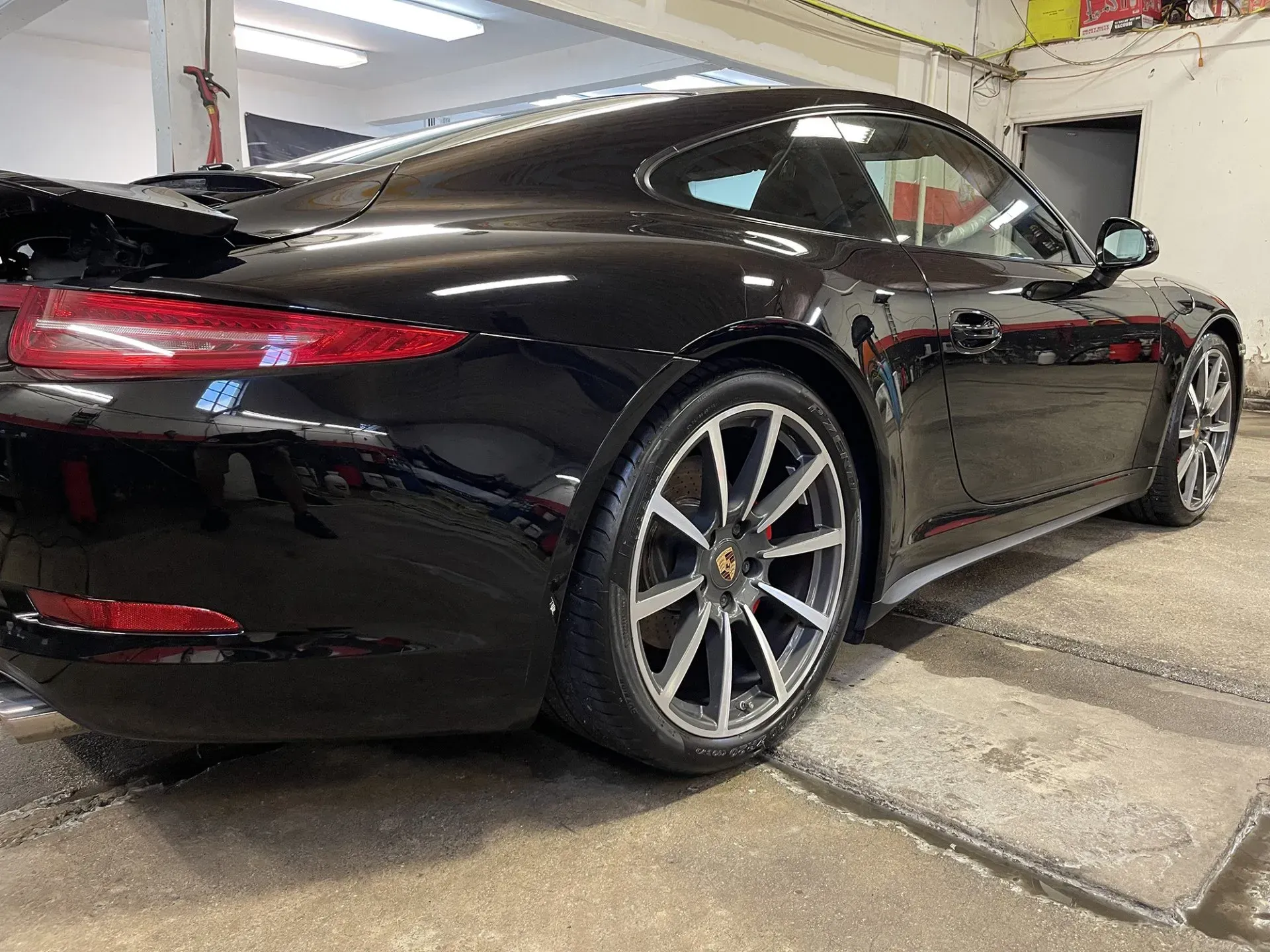Black Porsche sports car in a garage, rear view. Silver wheels, spoiler, and glossy finish.