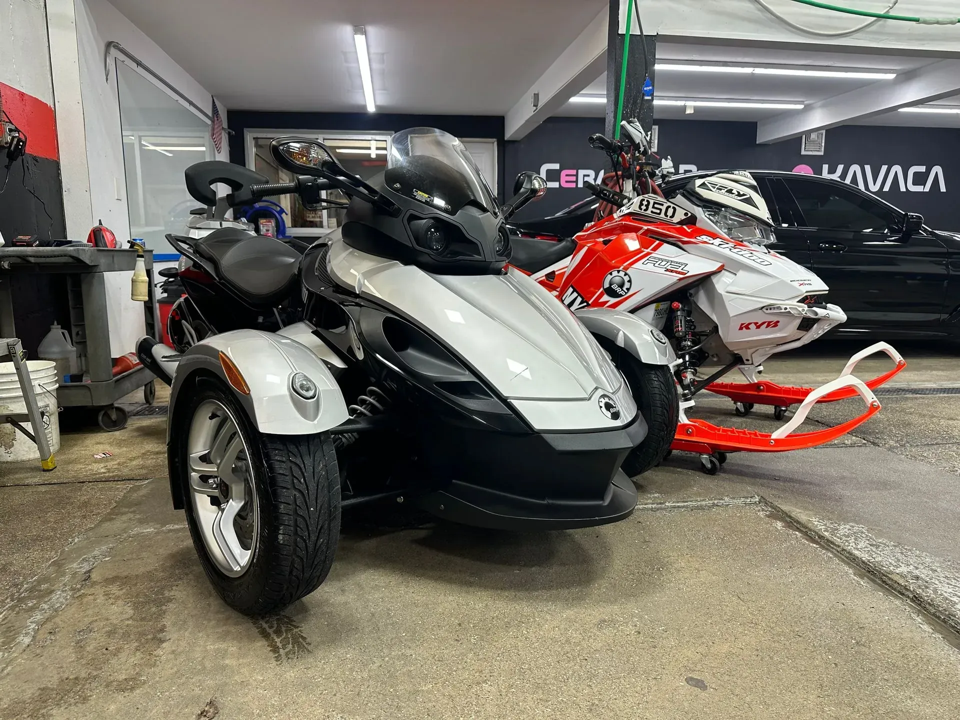 A white and black Can-Am Spyder three-wheeled motorcycle parked next to a snowmobile indoors.