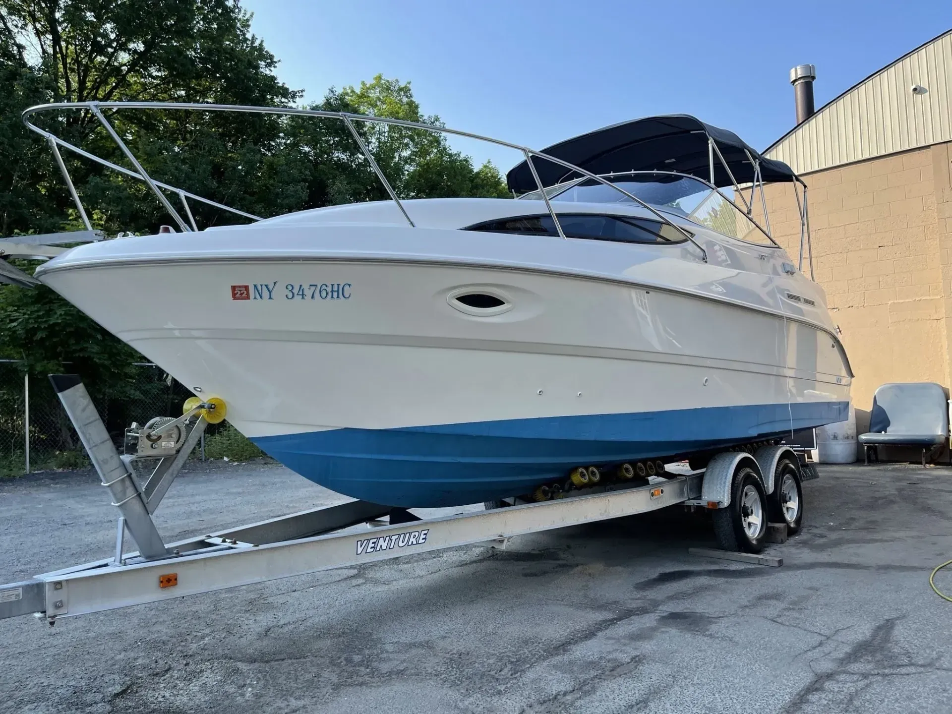 White and blue motorboat on a trailer, parked in front of a building, under a bright sky.