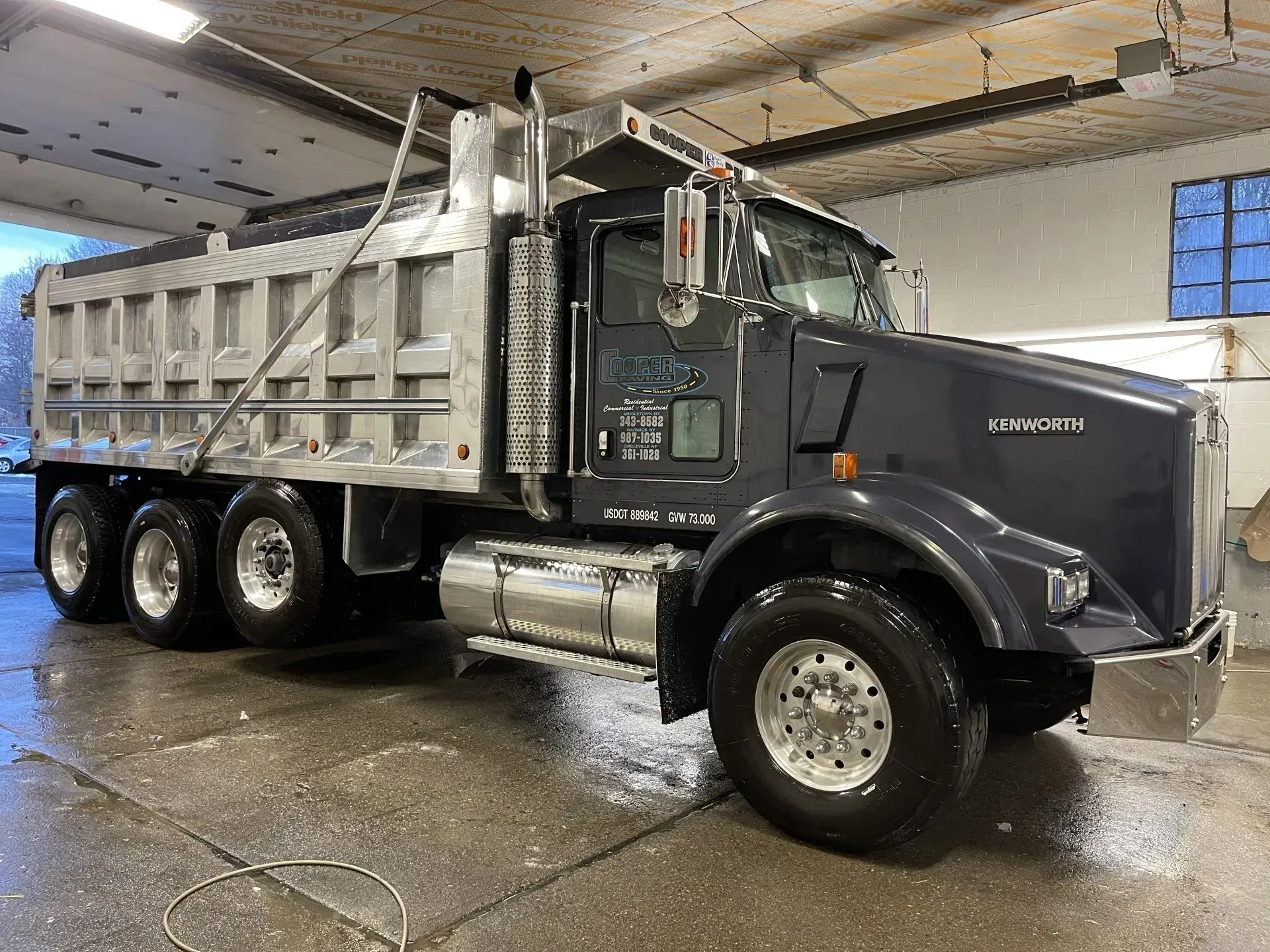 Dark gray dump truck, parked indoors, silver dump bed, wet surface, Kenworth logo on the door.