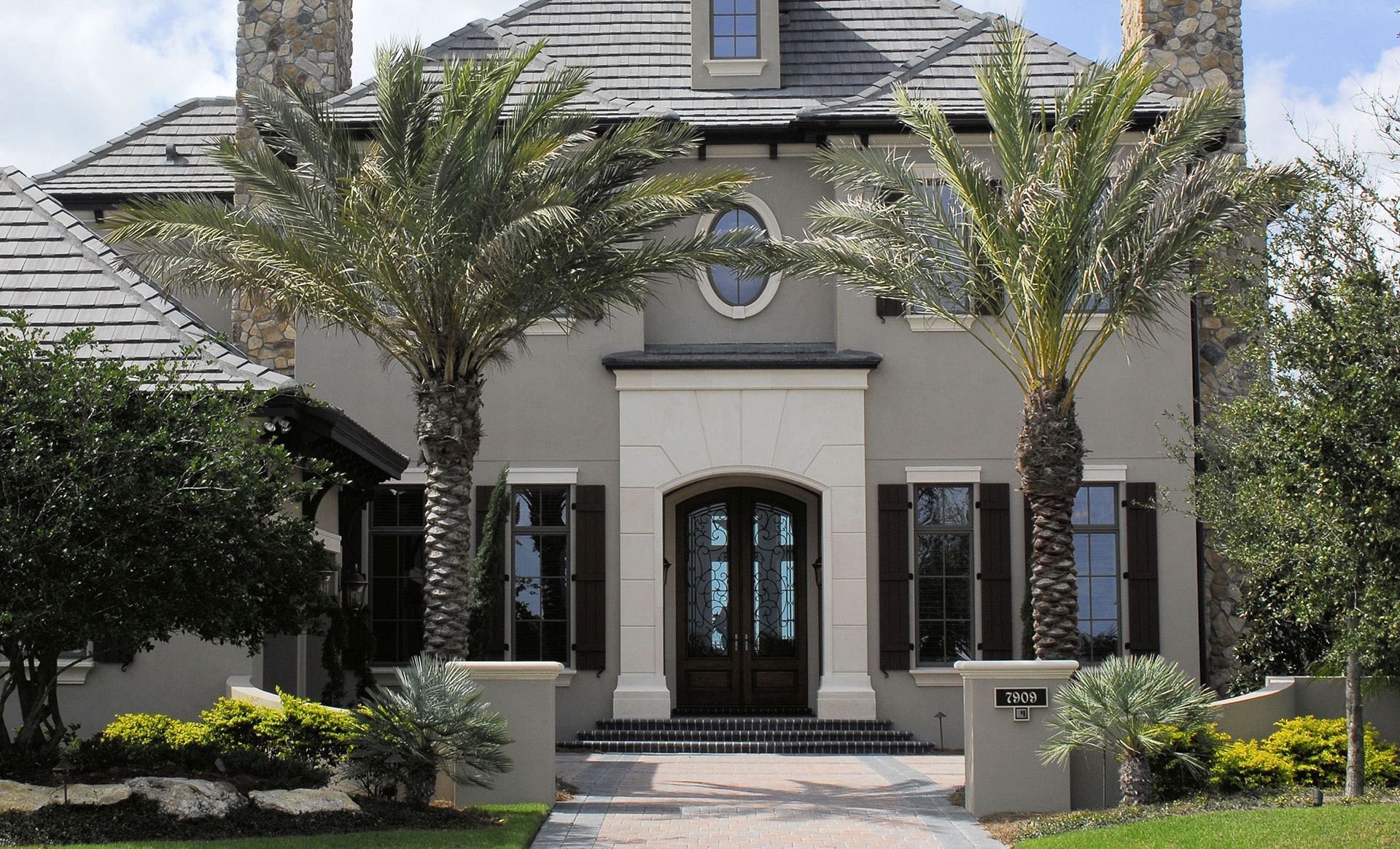 Beige house with a dark roof, palm trees, and a brick pathway.