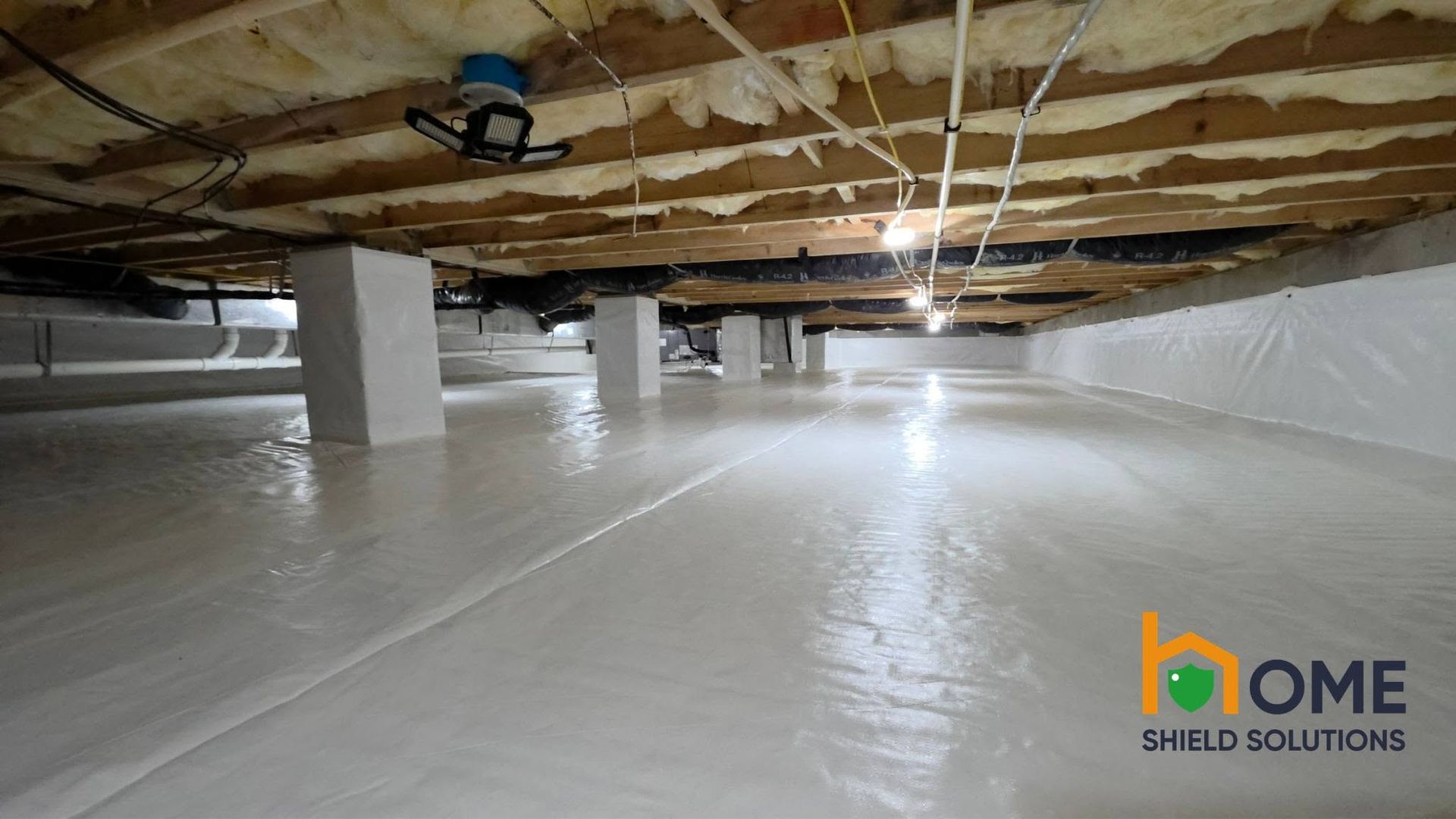 Crawlspace with white vapor barrier, exposed joists, support posts, and a Home Shield Solutions logo