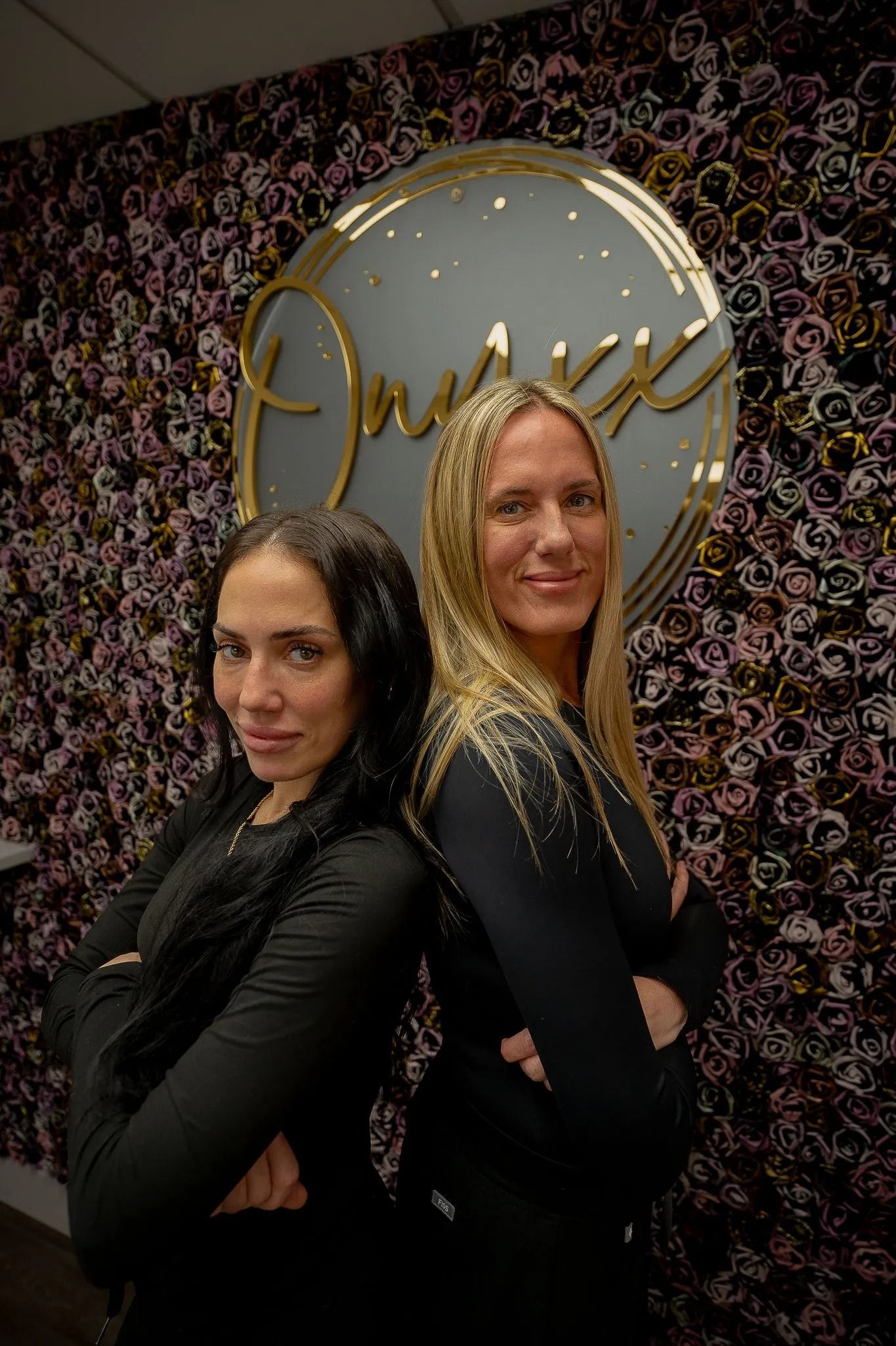 Two women in black tops stand back-to-back, arms crossed, in front of a logo on a patterned wall.