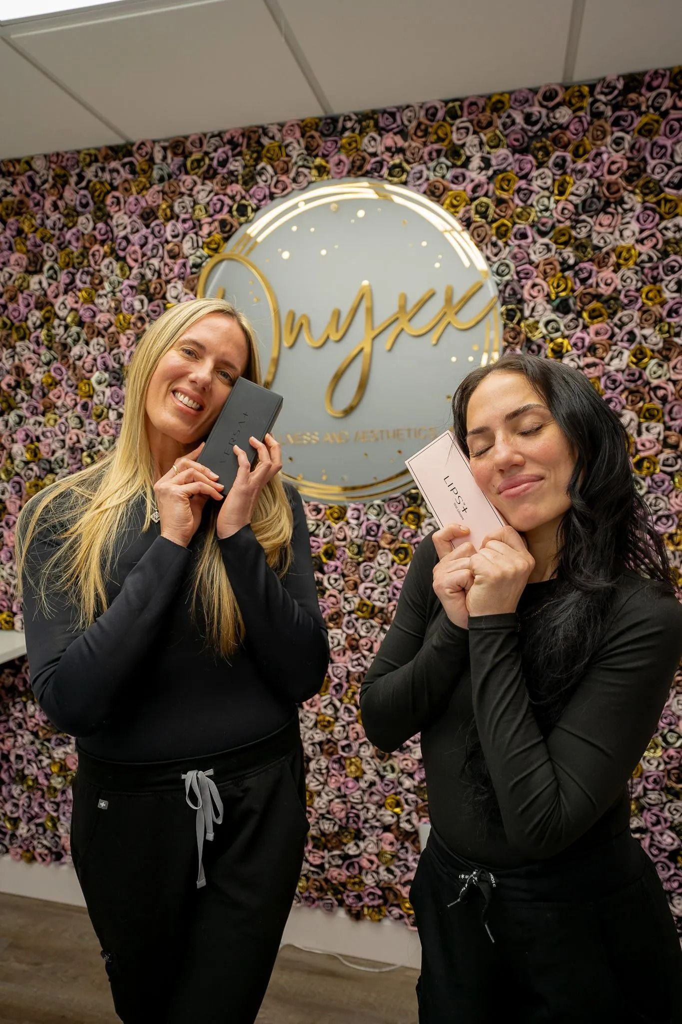Two women smiling, holding phones near their faces. Black outfits, pink and gold sign in background.