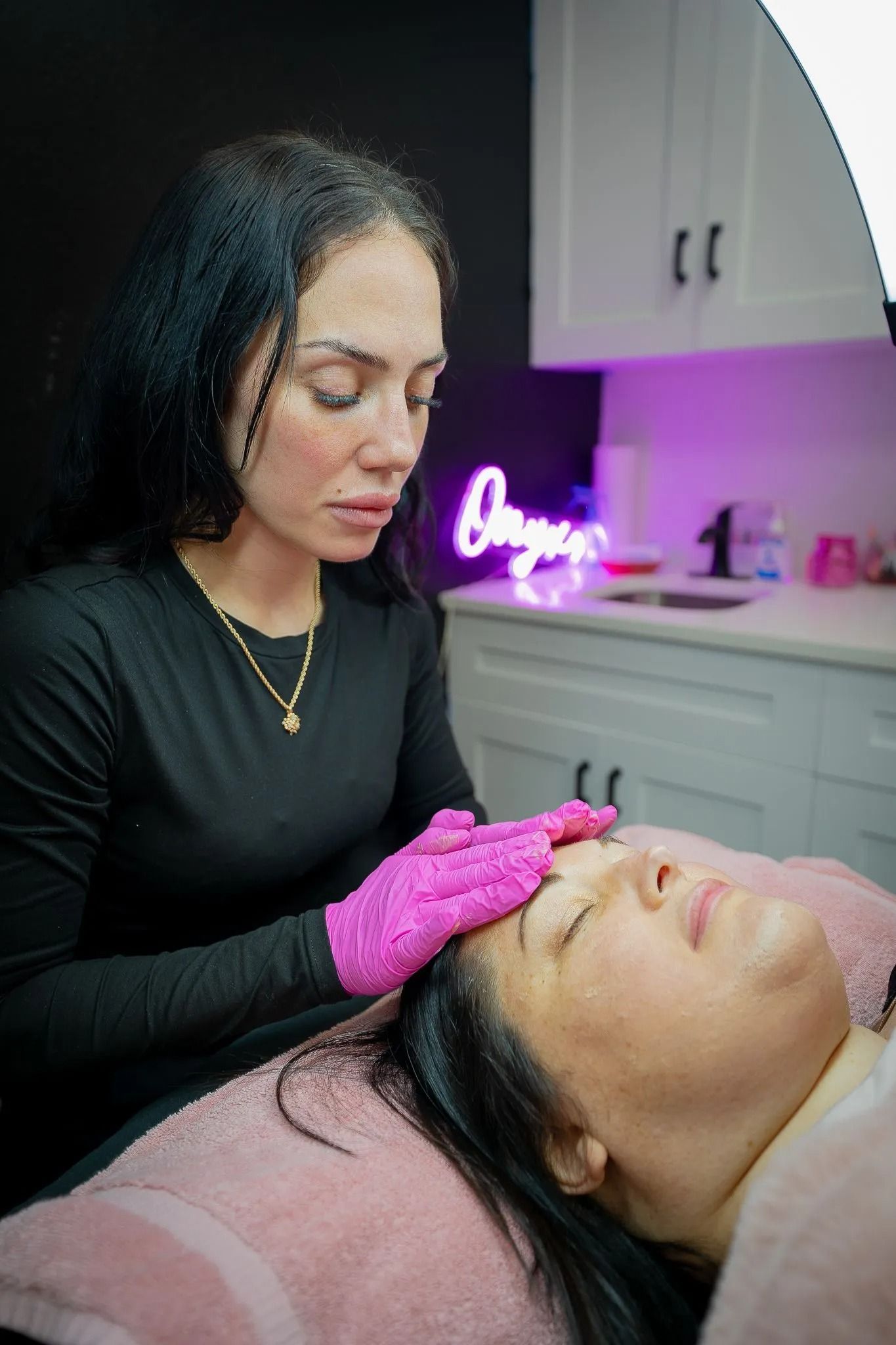 Woman in pink gloves performing a facial treatment on a client lying on a pink bed; bright salon.
