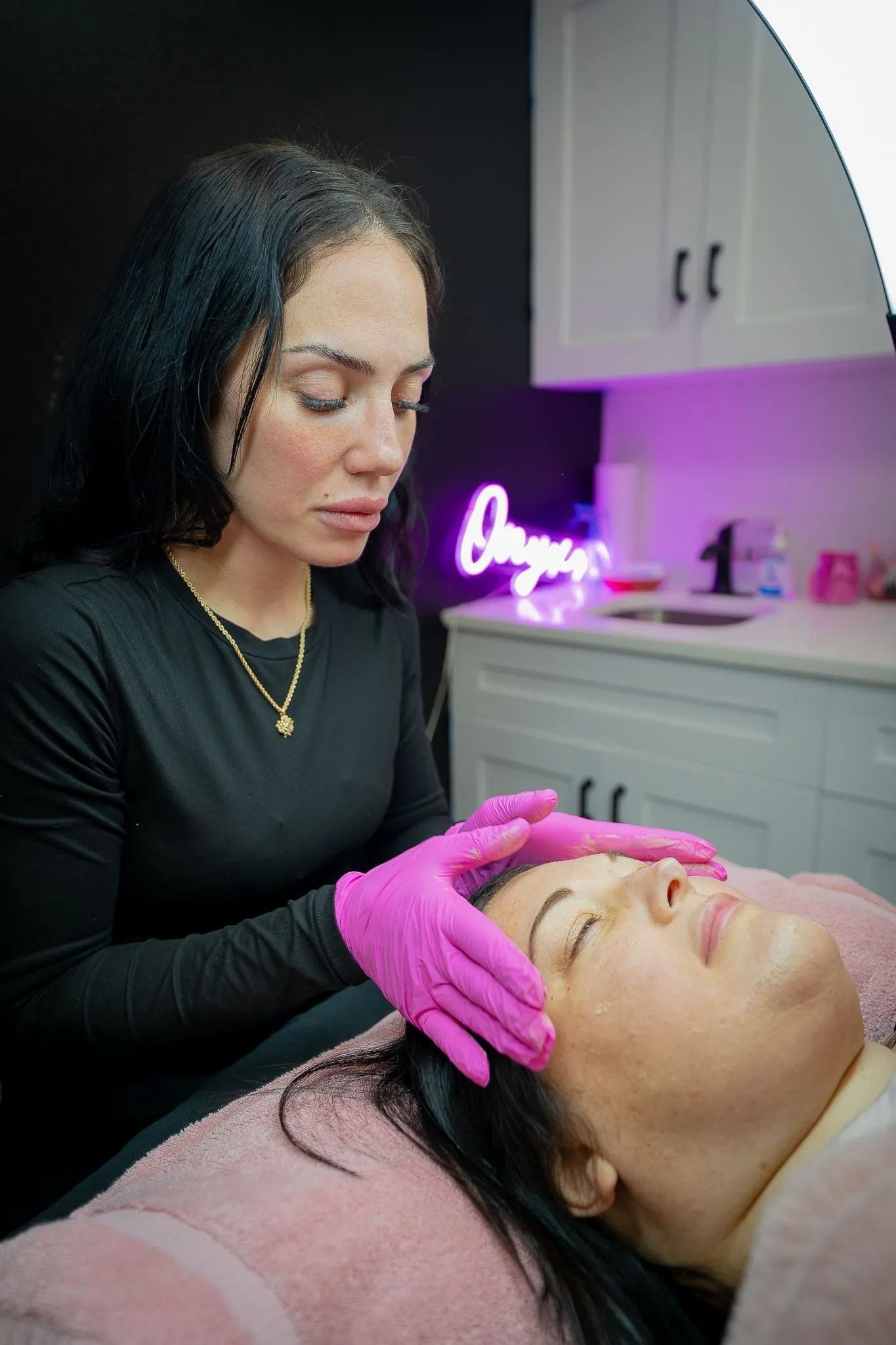 Woman performing a facial treatment on another woman in a spa setting. Both women have dark hair; one is wearing gloves.