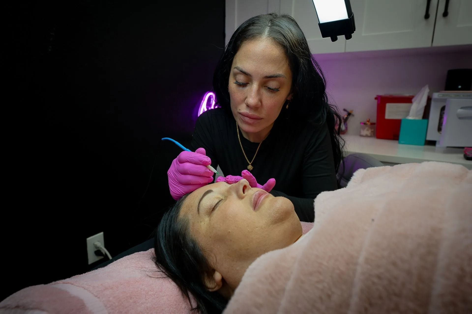 Woman getting eyebrow microblading; technician works on her eyebrow, black background, pink gloves.