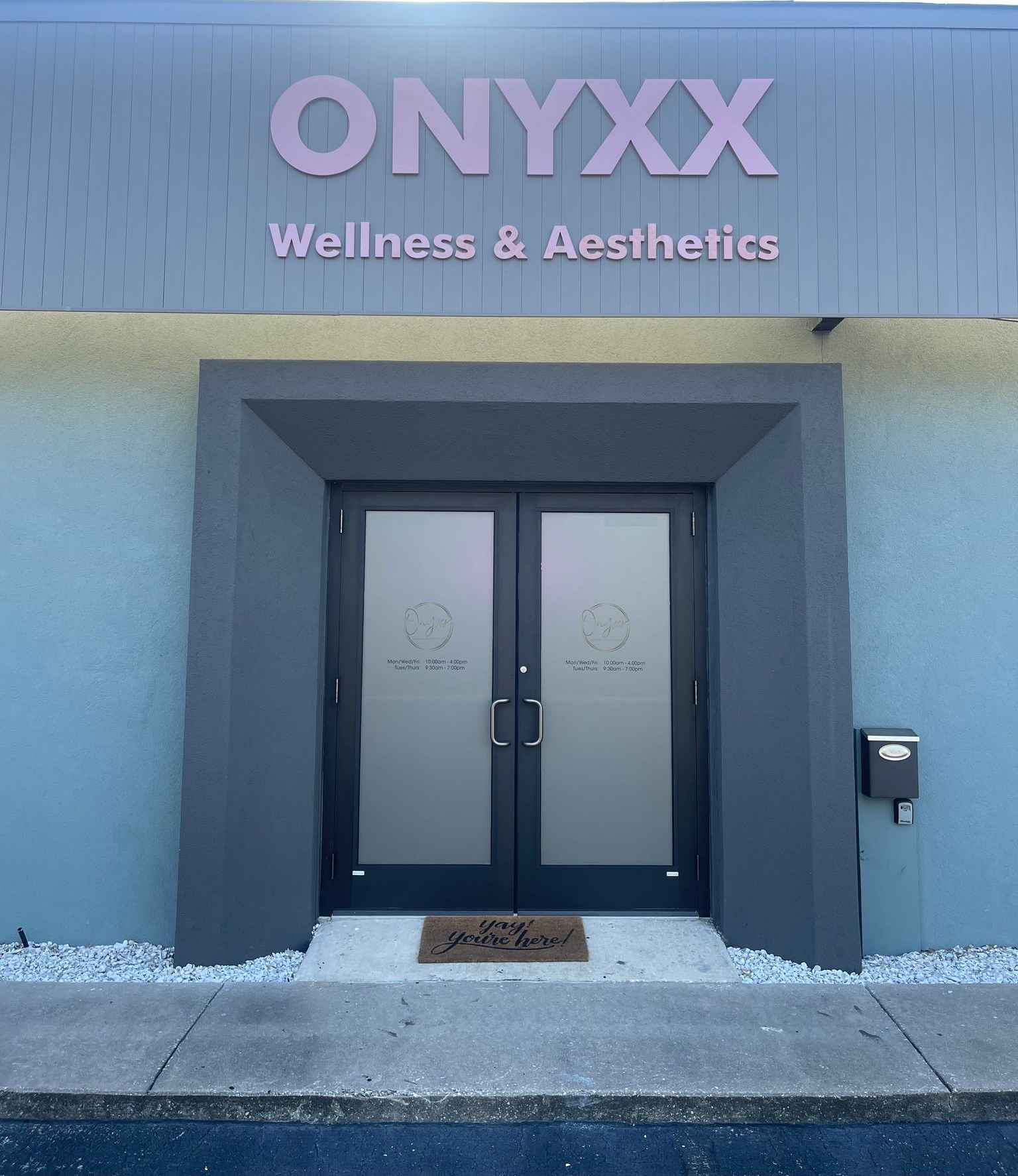 Exterior of Onyxx Wellness & Aesthetics building. Pink sign over gray entryway with double doors; a welcome mat.
