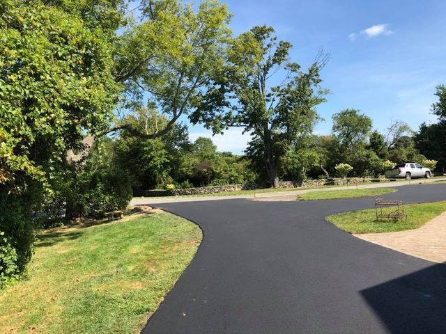A black asphalt driveway surrounded by trees and grass on a sunny day.