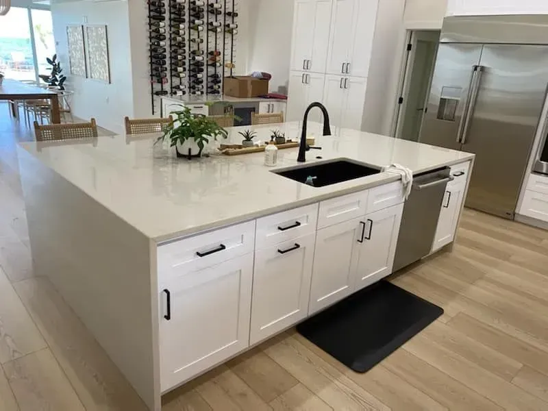 White kitchen island with black sink and faucet, dishwasher, and white cabinets. Light wood floor.
