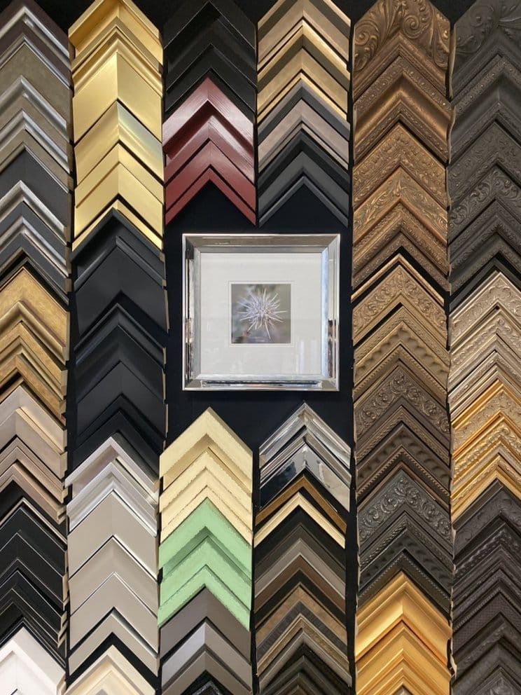 Display of various picture frame samples surrounding a framed artwork; diverse colors and styles.