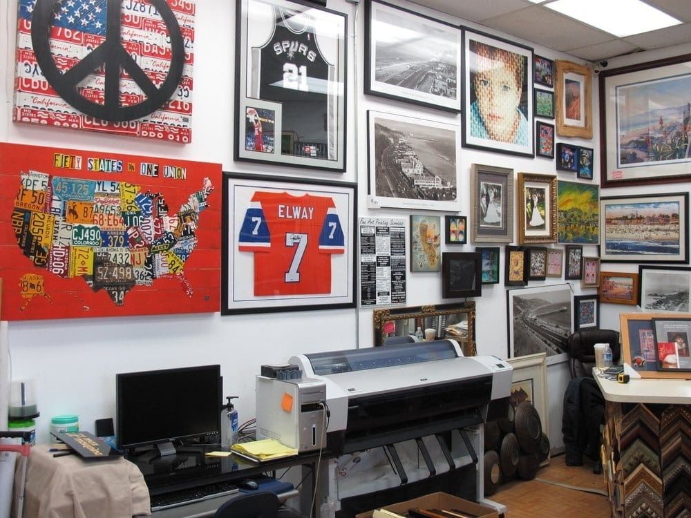 Interior of a frame shop. Various framed art and items displayed on walls. Workstation with computer and printer.