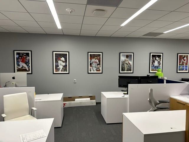 Office space with white desks, gray walls with framed sports posters, and overhead lights.