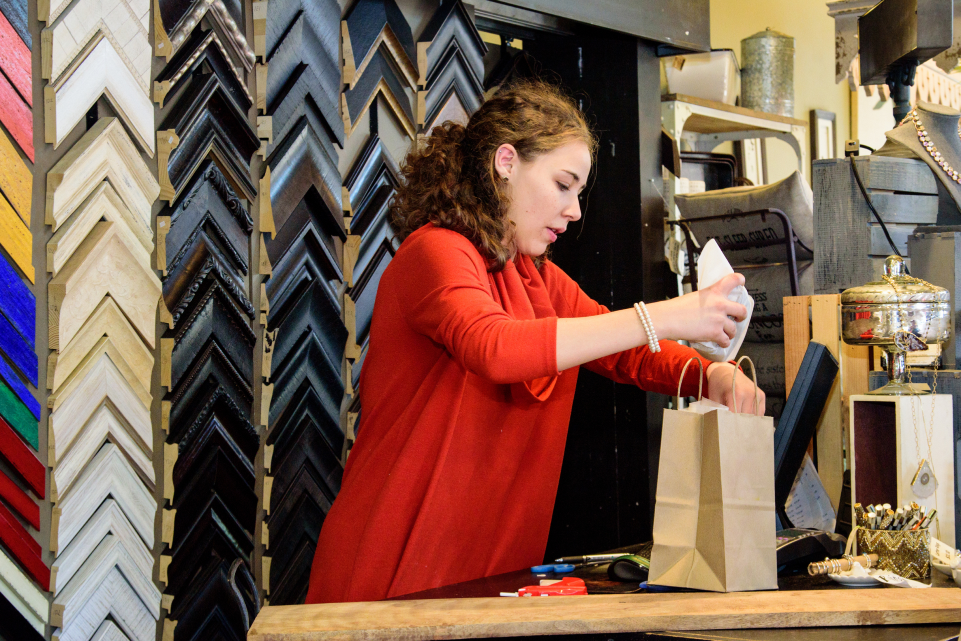 Woman in red shirt packaging something at a frame shop counter with frame samples in the background.