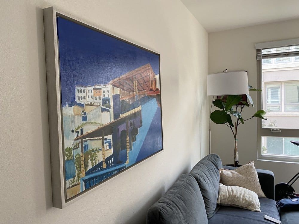 Large painting on wall, depicting a cityscape in blue and white tones. Grey-framed, above a dark couch.