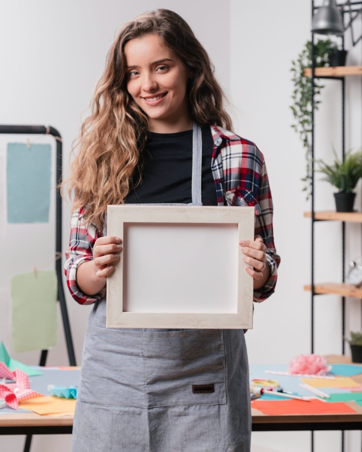 Woman holding a blank, white framed picture, smiling in a workshop with craft supplies.