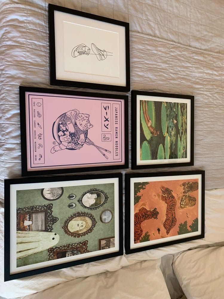 Five framed art pieces with black frames on a bed. Styles range from line drawings to illustrations and abstract art.