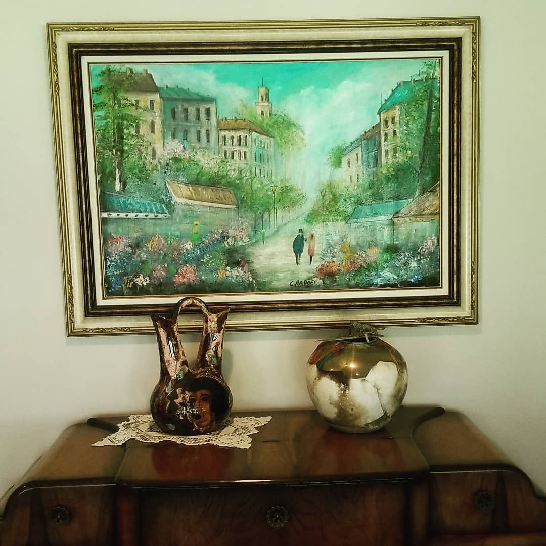 Painting of a street scene, framed, above a wooden cabinet. Two vases sit on the cabinet.