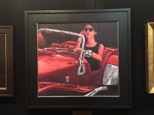 Painting of a person in sunglasses driving a red convertible, framed on a dark wall.