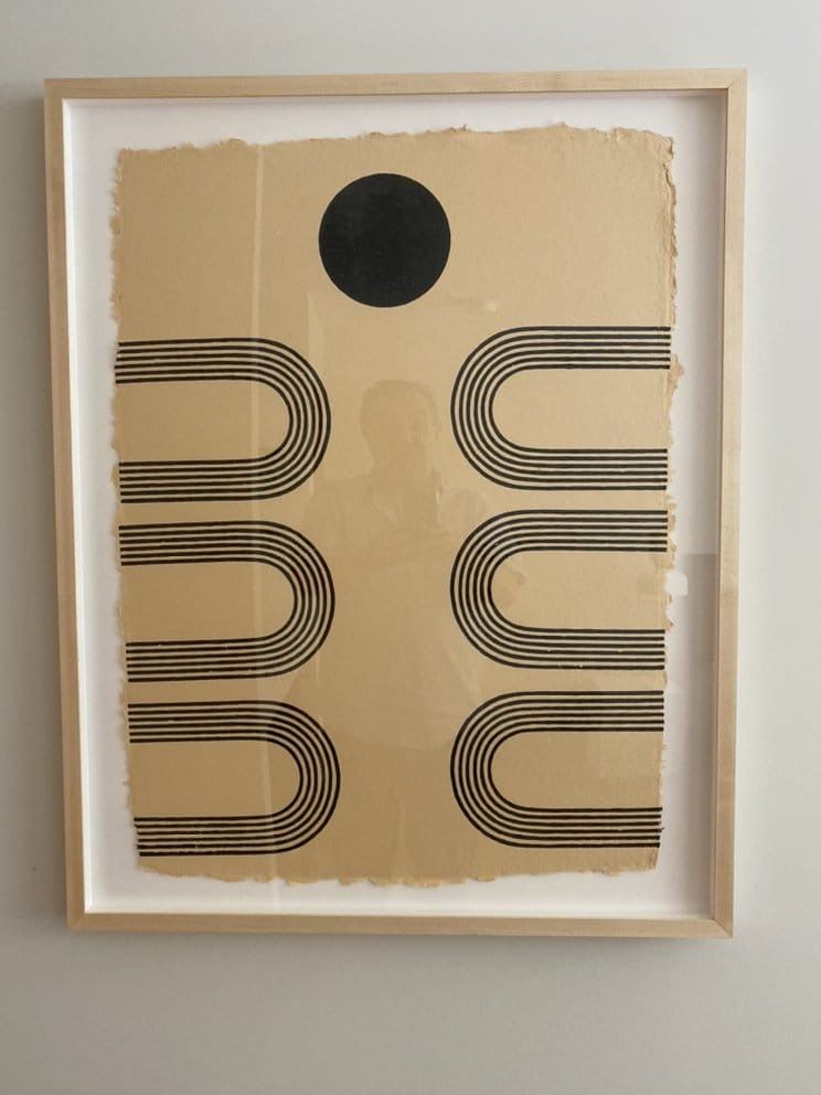 Abstract artwork: Black shapes on tan paper, framed. Black circle at top, curved lines below.