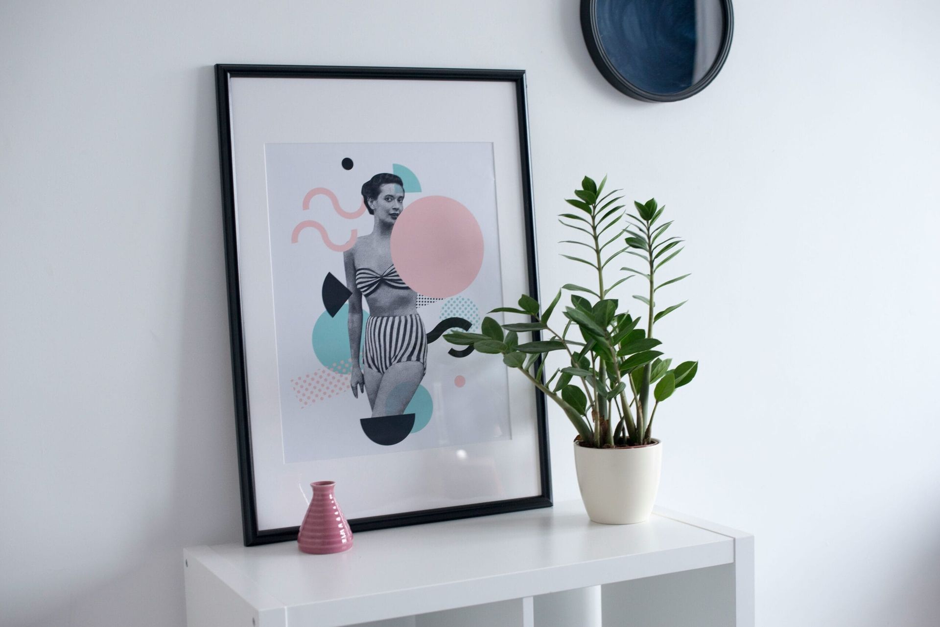 A framed art print of a person with abstract shapes, a potted plant, and a pink vase on a white shelf.