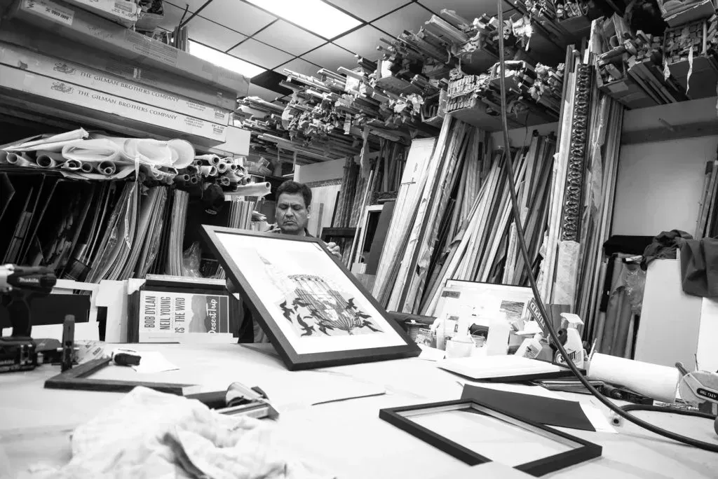 Man in a frame shop holding framed artwork; surrounded by frames and materials on a workbench.