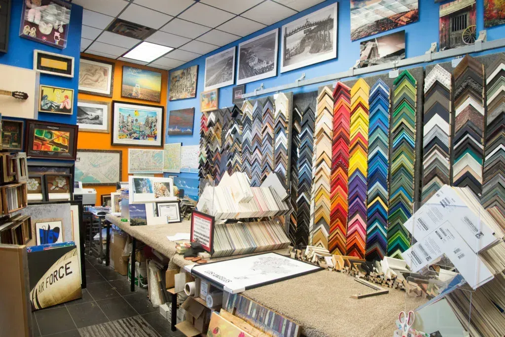 Interior of a framing shop; artwork and frame samples displayed on walls and work surfaces.