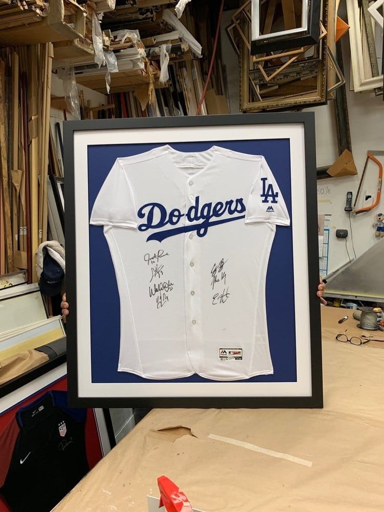Framed Los Angeles Dodgers jersey with signatures, displayed on a blue background, in a framing shop.