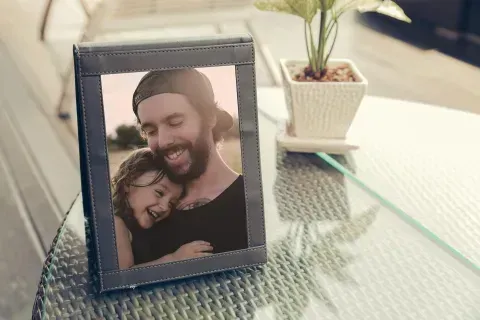 Framed photo of a man hugging a child; both smiling. The frame is on a glass table next to a small potted plant.