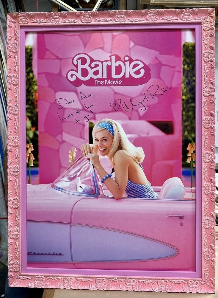 Poster of Margot Robbie in a pink Barbie movie car, framed with a pink carved border.